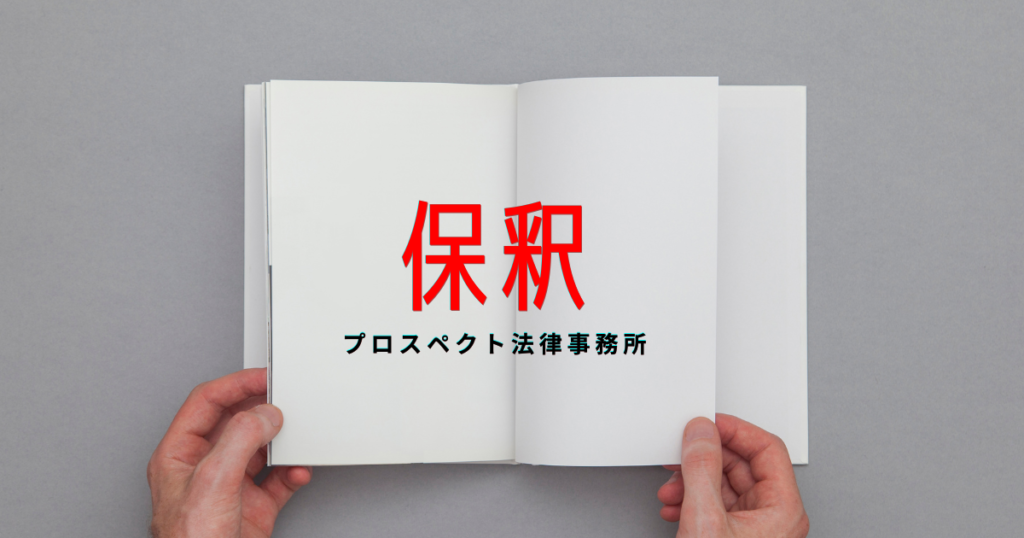 相談室の机に置かれた書類と封筒、時計を前に座る人物の手元